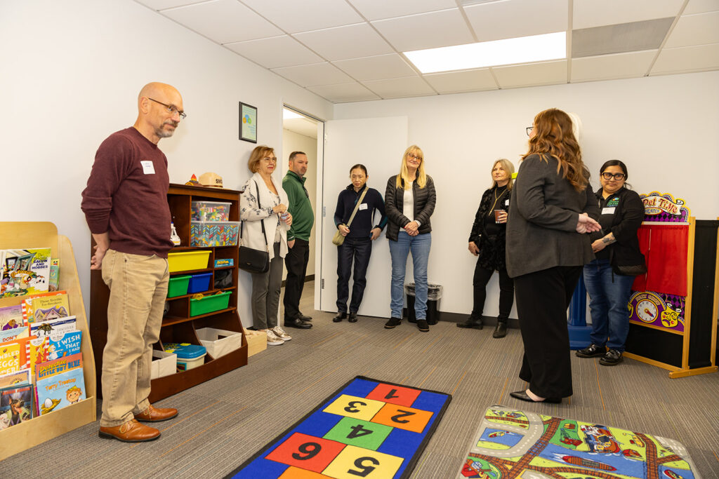 A group of guests listen as a tour guide discusses the San Pedro play therapy room, which is filled with children's books, toys, and colorful rugs designed to create a comforting and engaging space for young clients.